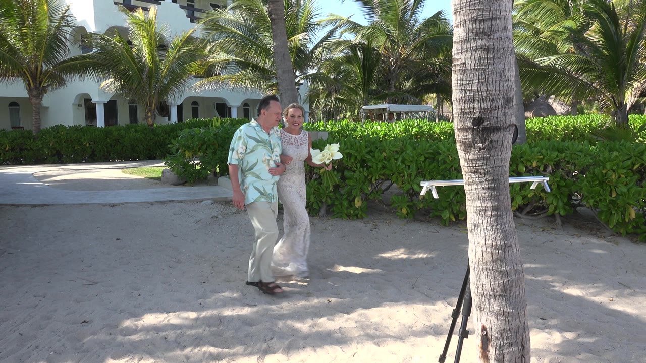 At 0:02 in Playa Del Carmen, the father and bride pass near a large palm trunk, with the bride's bouquet visible against the green hedge line.