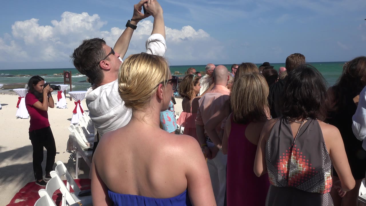 Guests stand on a beach facing a wedding ceremony by the ocean.