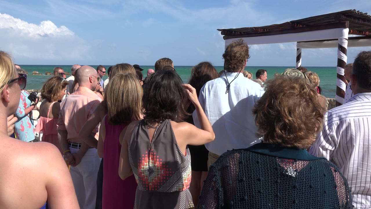 Wedding guests seen from behind facing a beach ceremony canopy by the ocean.