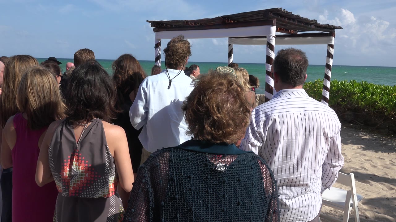 Wedding guests seen from behind face a striped canopy on the sand in Playa Del Carmen, Mexico, with turquoise water and bright sky beyond.