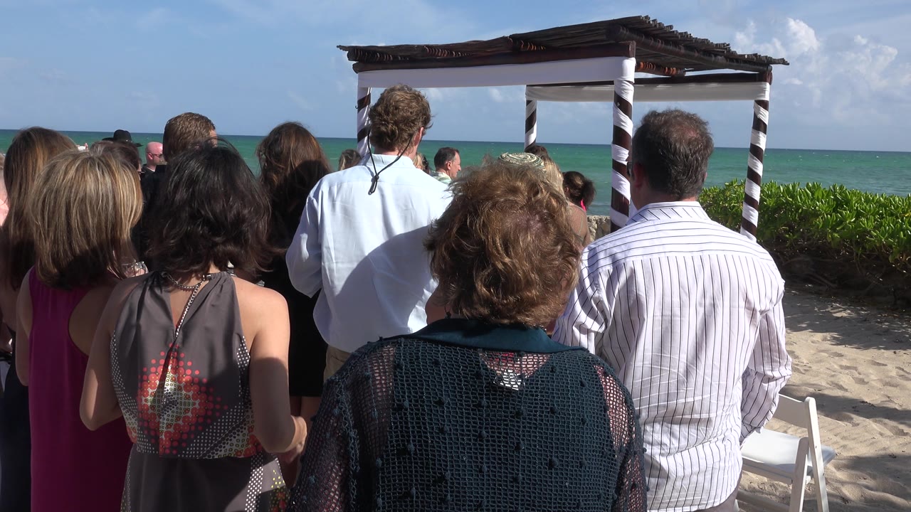 Beach wedding guests look toward the ceremony canopy and sea.