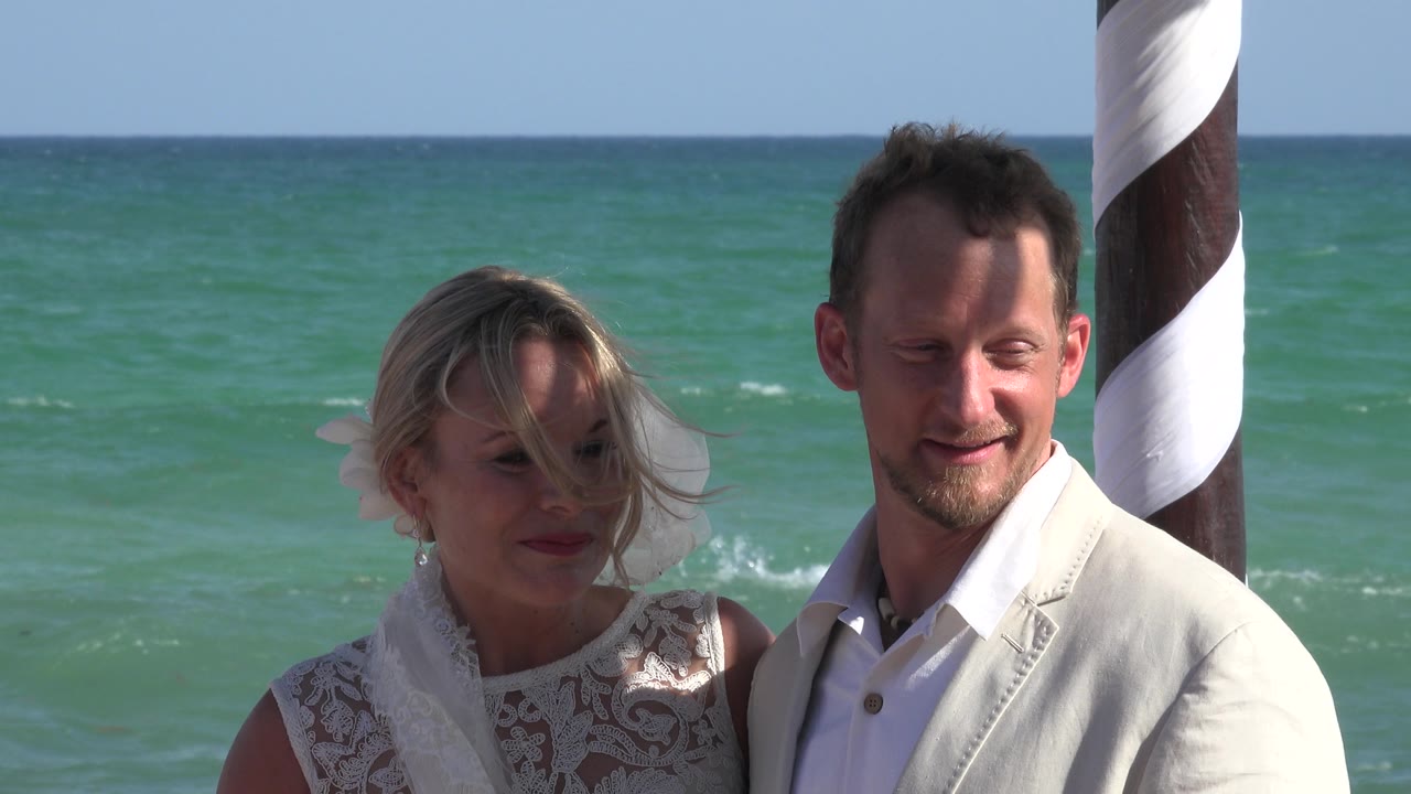 At 0:00, a couple stands close together in Playa del Carmen with turquoise ocean water behind them and a brown-and-white striped pole at the right edge.
