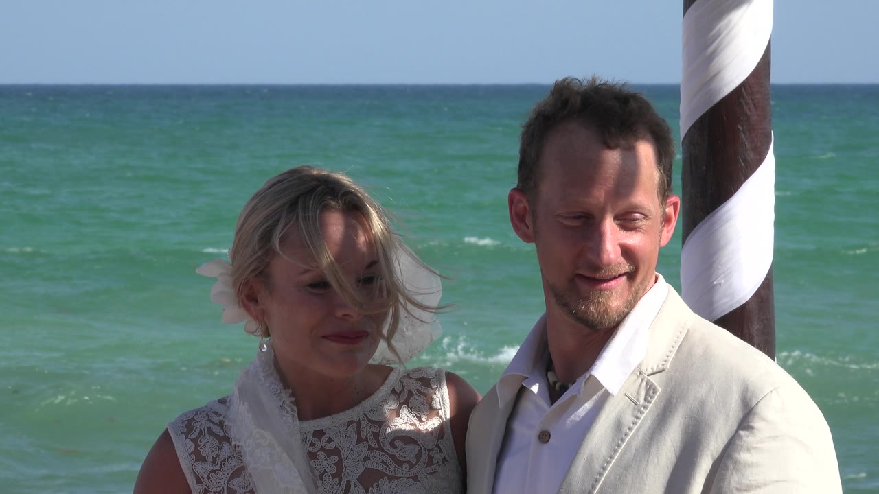 At 0:01, the couple remains in a tight beach portrait in Playa del Carmen as wind blows the woman's hair across her face and the ocean fills the background.