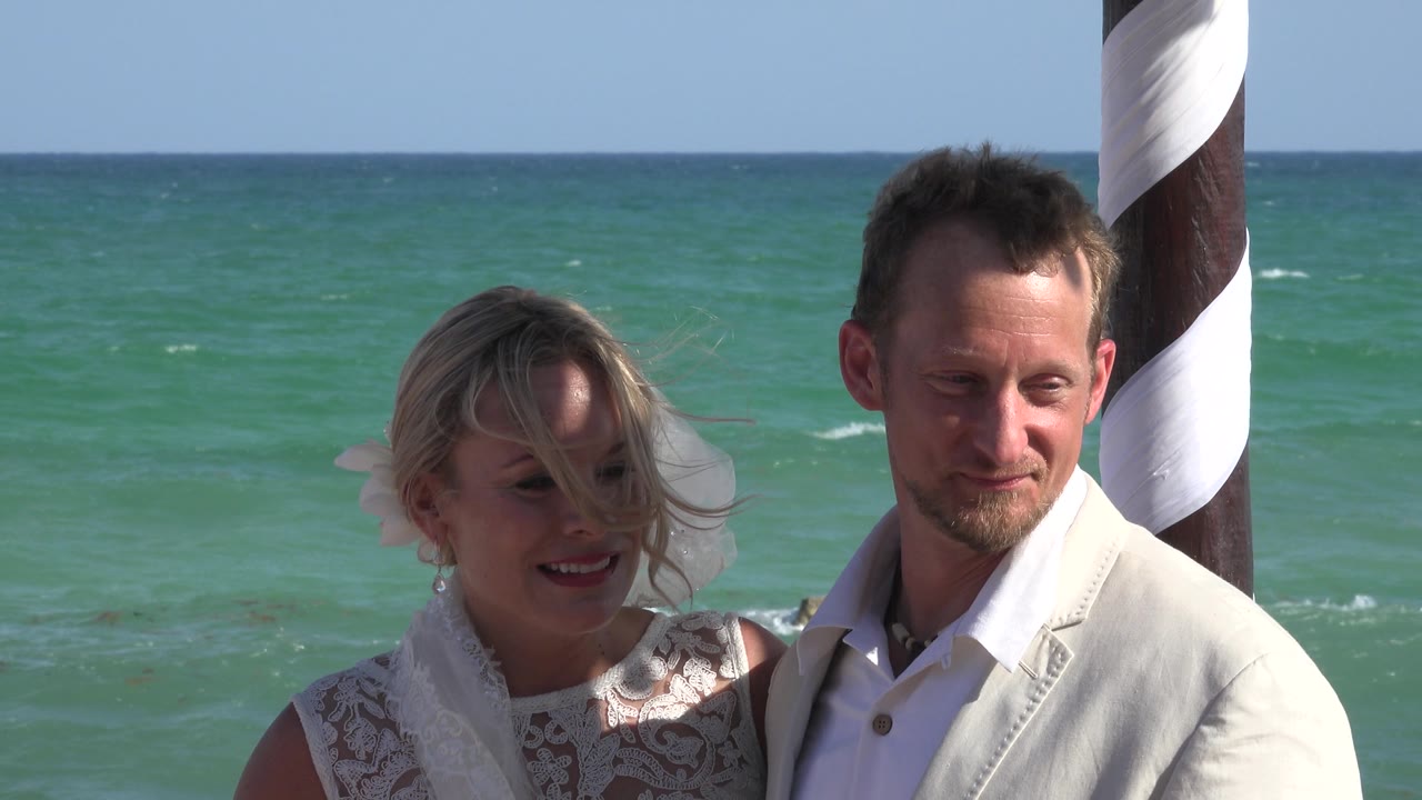Smiling couple near turquoise water with striped pole at right.
