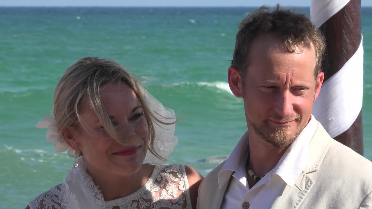 At 0:01, the woman looks toward the man as wind moves hair across her face, while the man faces forward near the striped pole in Playa del Carmen, Mexico.
