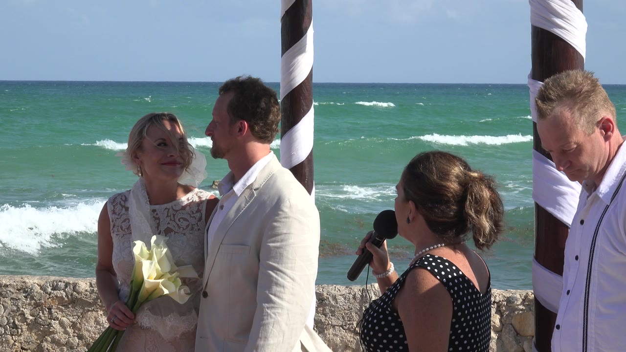 At Playa del Carmen, Mexico, a bride holding white flowers stands beside the groom while an officiant with a microphone faces them near the ocean.