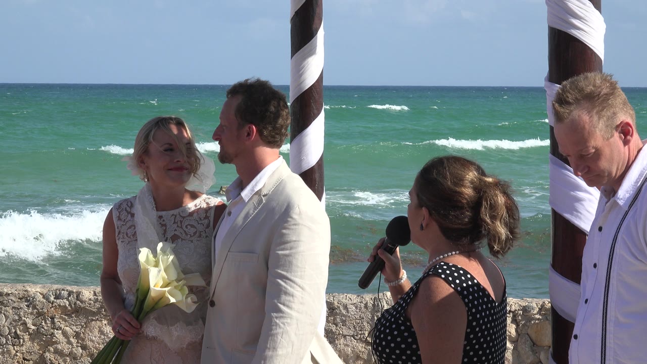 At Playa del Carmen, Mexico, the bride and groom stand close together beside the seawall as the officiant speaks with the turquoise ocean behind them.