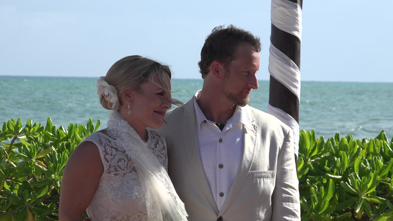 The bride and groom remain framed near the striped ceremony post in Playa del Carmen, with the groom looking downward and the ocean horizon running behind the green hedge.