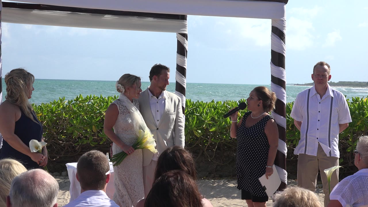 Beach wedding ceremony with couple, officiant, attendants, and seated guests by the ocean.