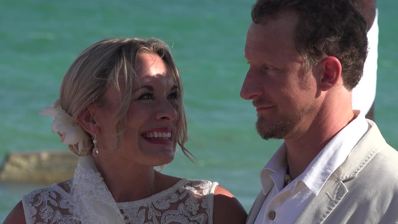 In Playa Del Carmen, Mexico, a blonde bride in a white lace dress smiles up at a groom in a light suit with turquoise water behind them.