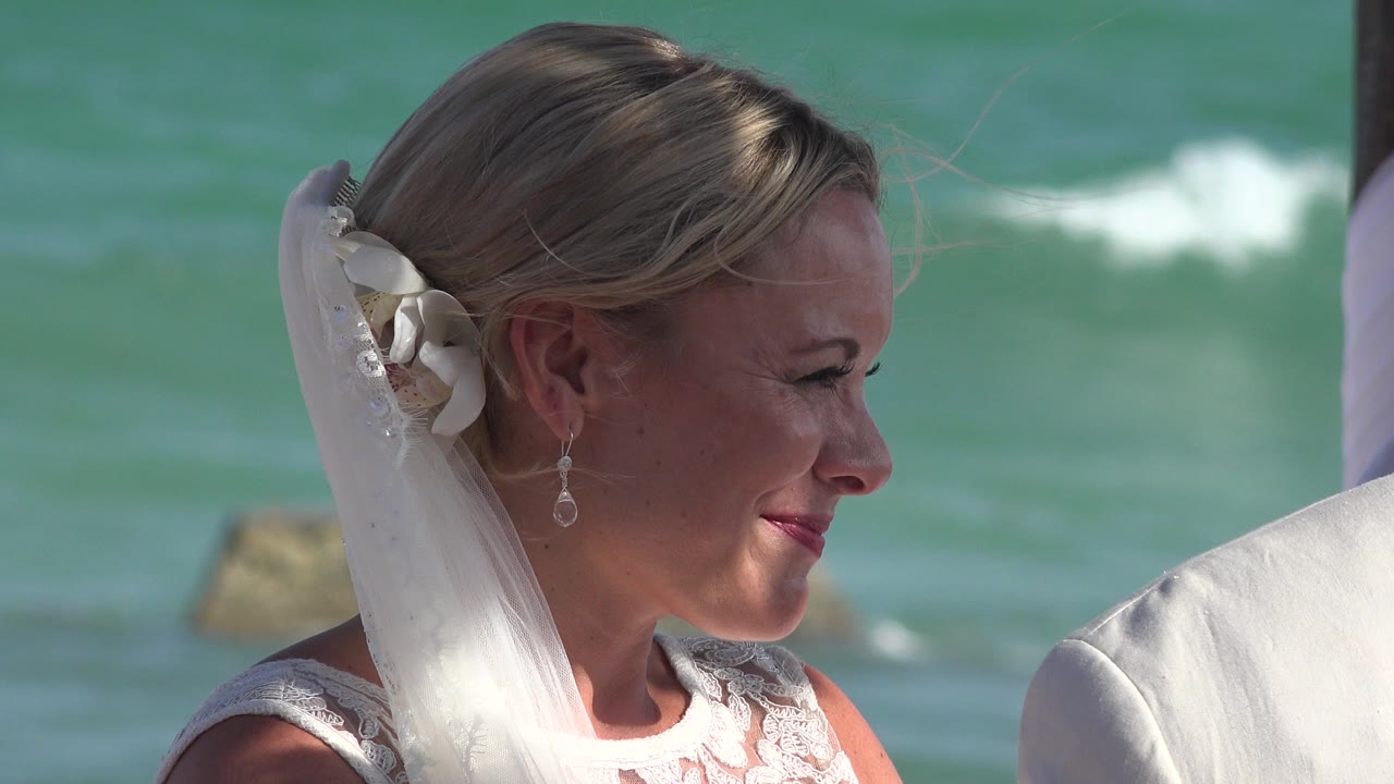 A blonde bride in Playa del Carmen smiles in side profile, with her veil and earring visible against turquoise ocean water.