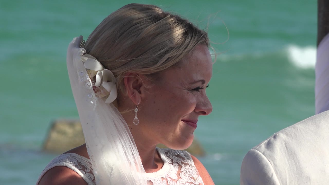 Bride in white veil smiles near the sea.