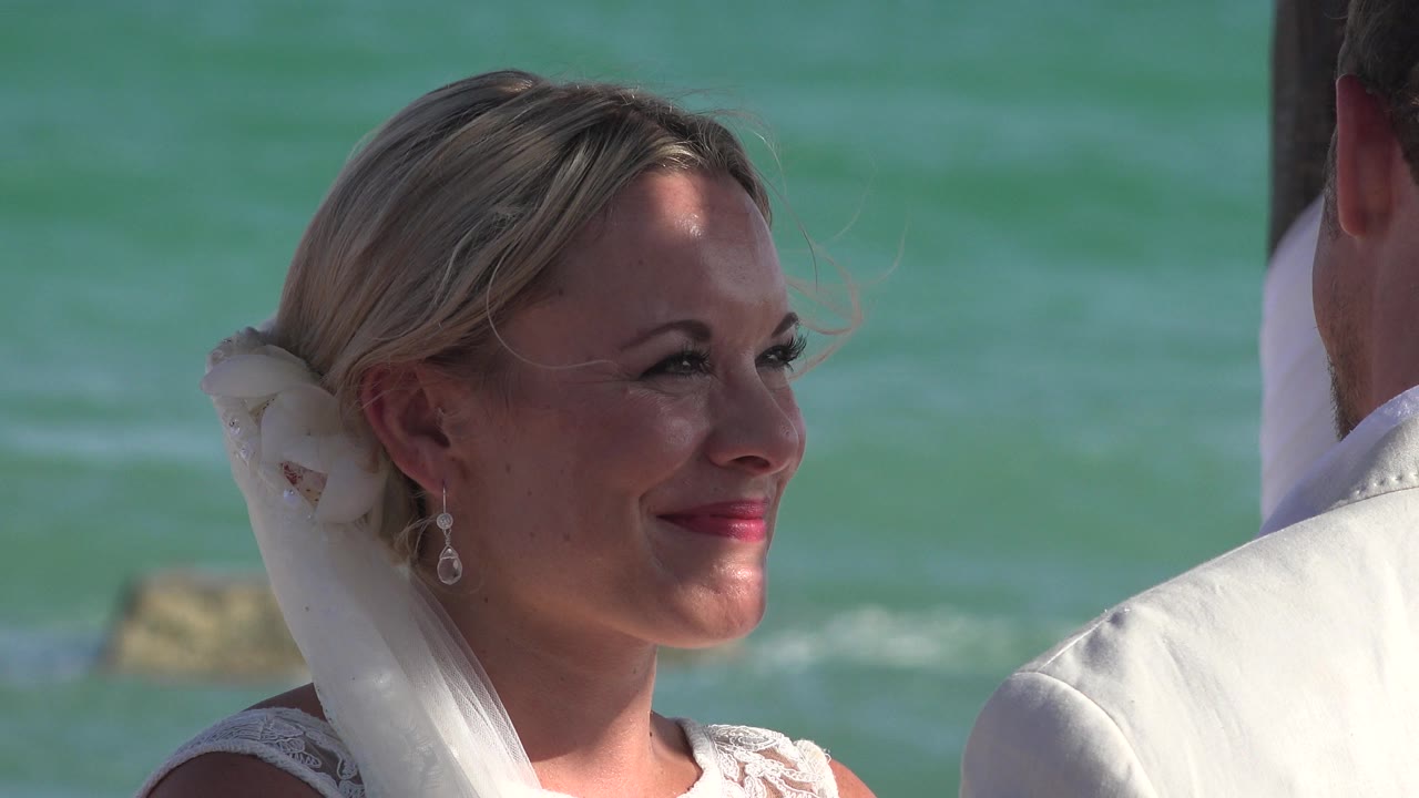 Bride smiling beside ocean water with a man partly visible at frame edge.