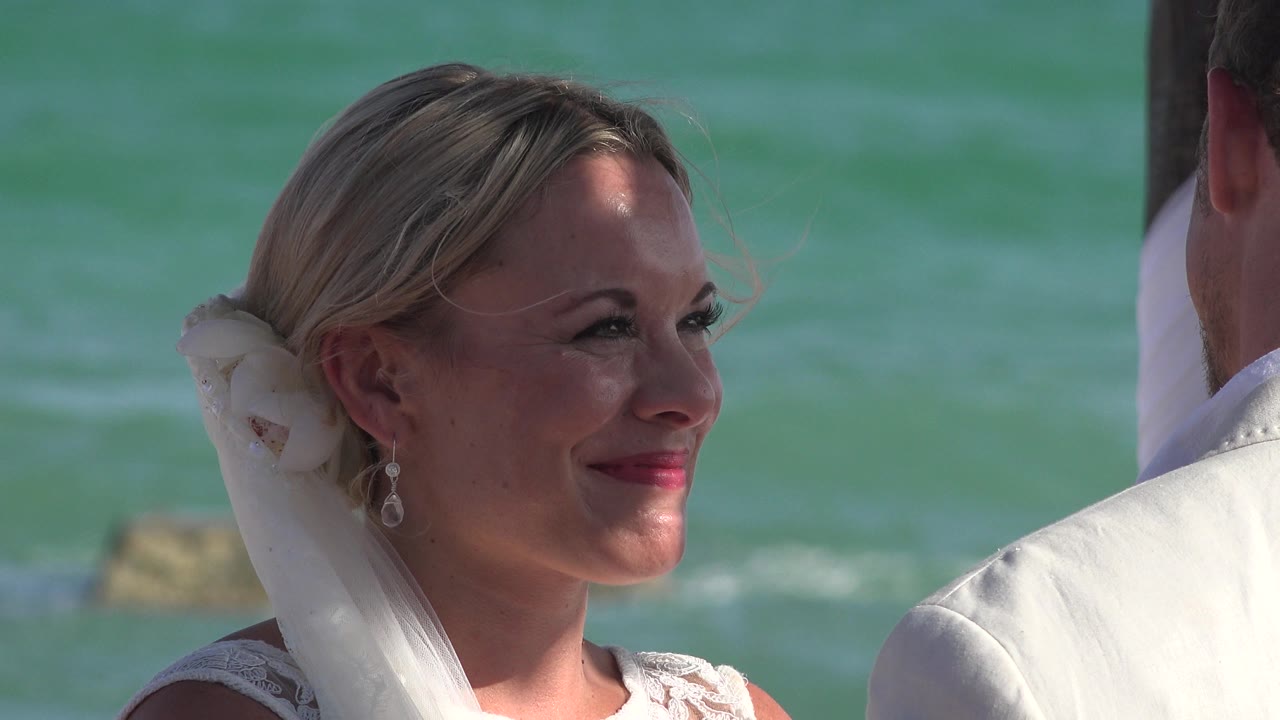 At Playa Del Carmen, Mexico, the bride keeps her face turned toward a man at the right edge while the ocean behind her appears teal with low waves and sunlit highlights.