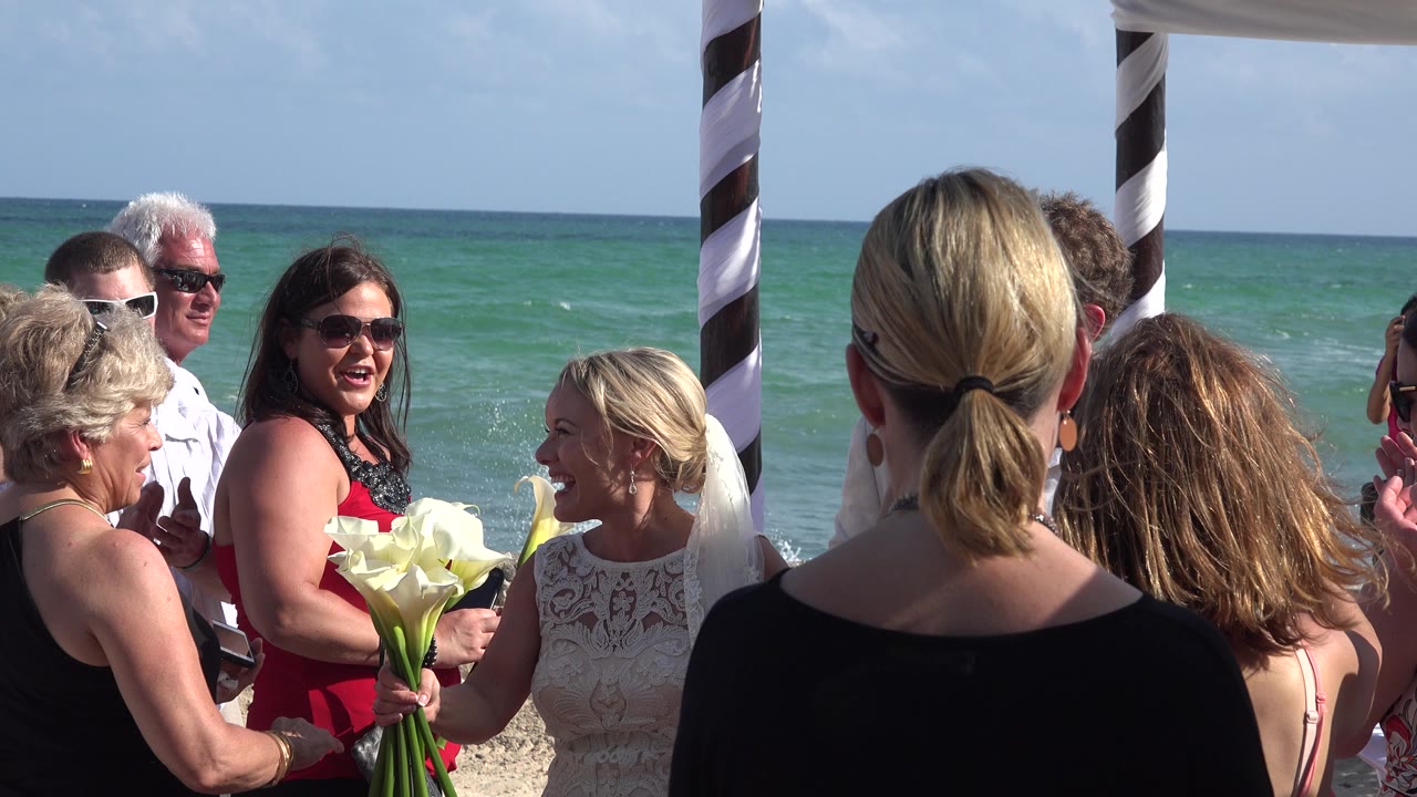 Bride holding calla lilies among wedding guests beside the ocean.