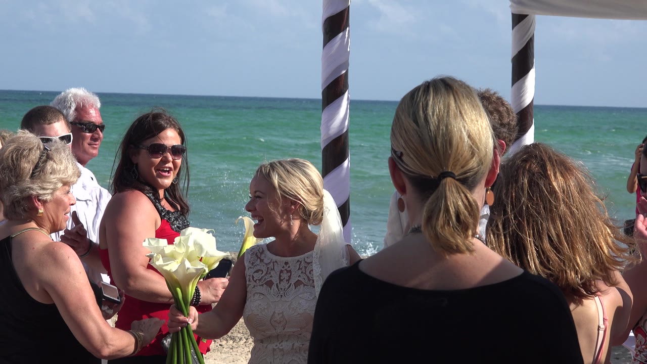 The bride smiles beside guests in bright sun while a striped ceremony post and the turquoise water mark the Playa Del Carmen beach setting.