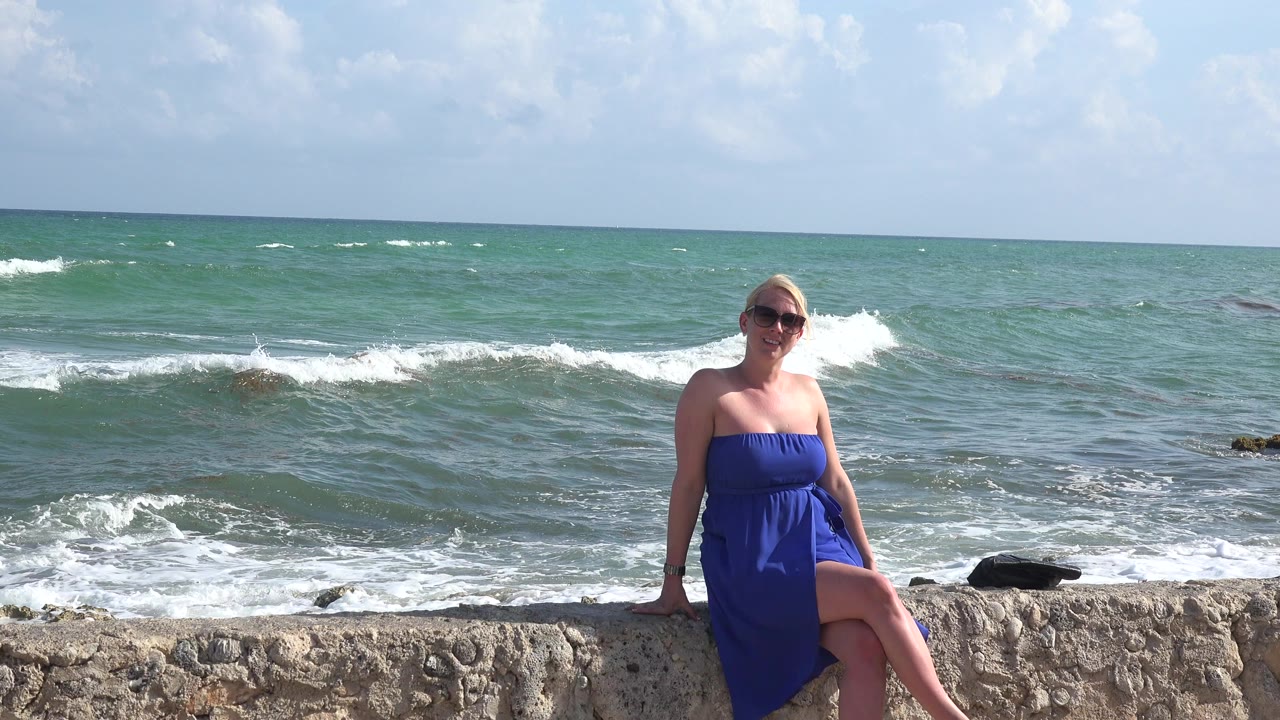 Woman in blue dress seated on a stone seawall beside ocean waves.