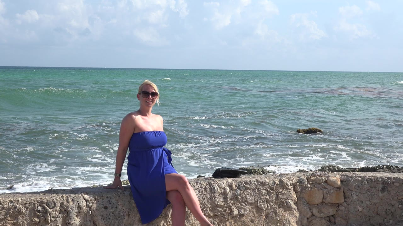 Woman in blue dress sitting on a stone wall by the ocean.