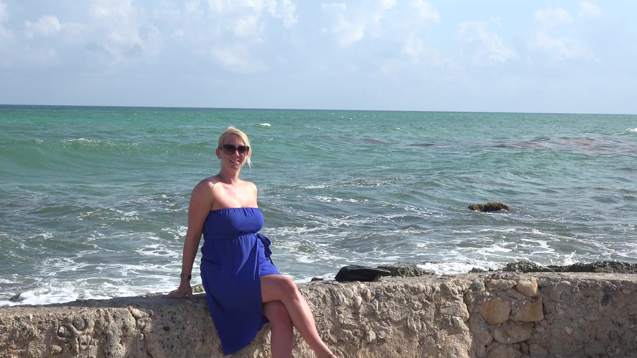 At 0:01, the woman remains seated on the stone wall as small whitecaps form across the Playa del Carmen ocean background.
