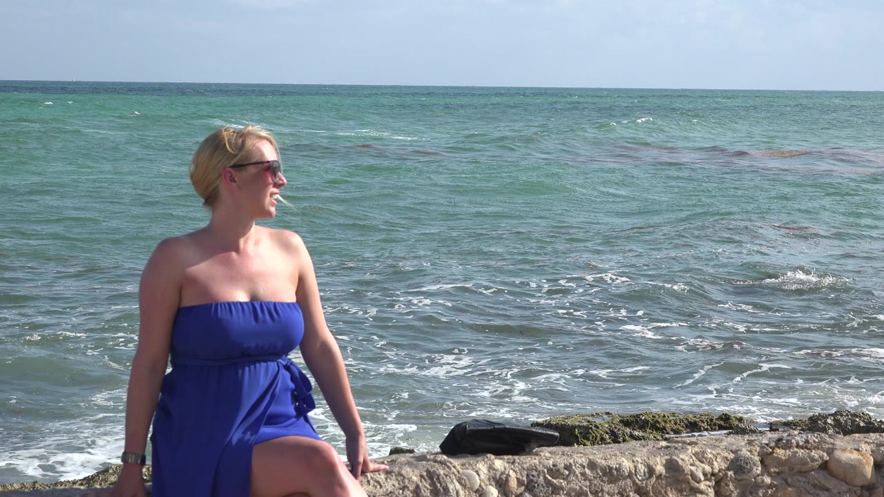 A woman in sunglasses and a blue strapless dress stands near a low stone edge with Playa del Carmen ocean water and a straight horizon behind her.