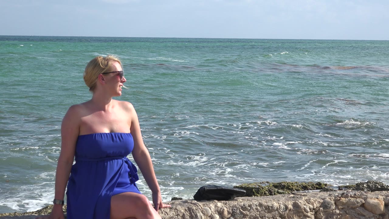 The woman remains near the left side of frame in Playa del Carmen, looking off camera while foamy waves move across the green-blue water behind her.