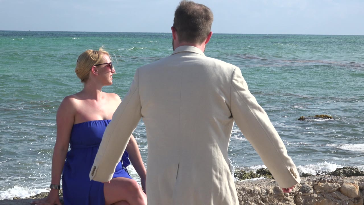 In Playa Del Carmen, Mexico, the woman looks toward the man as he stands with his back partly to camera, with low surf and dark patches in the water behind them.