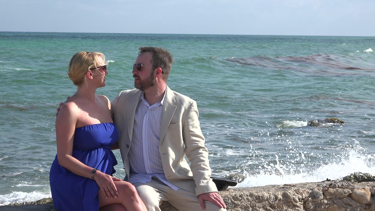 At 0:00, a woman in a blue dress and a man in a light suit sit close together on a stone edge in Playa Del Carmen, Mexico, with green-blue water and small surf behind them.