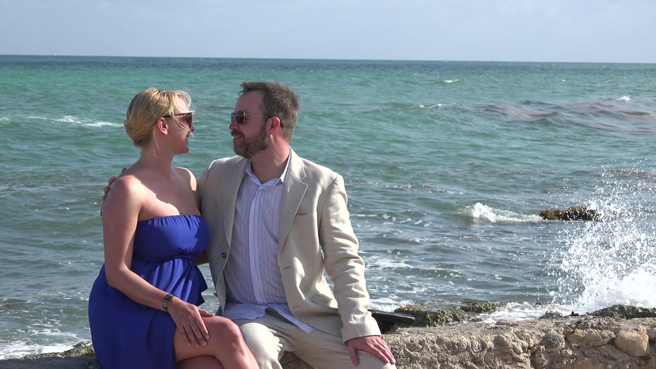 Couple seated on a stone edge with a wave splashing behind them.