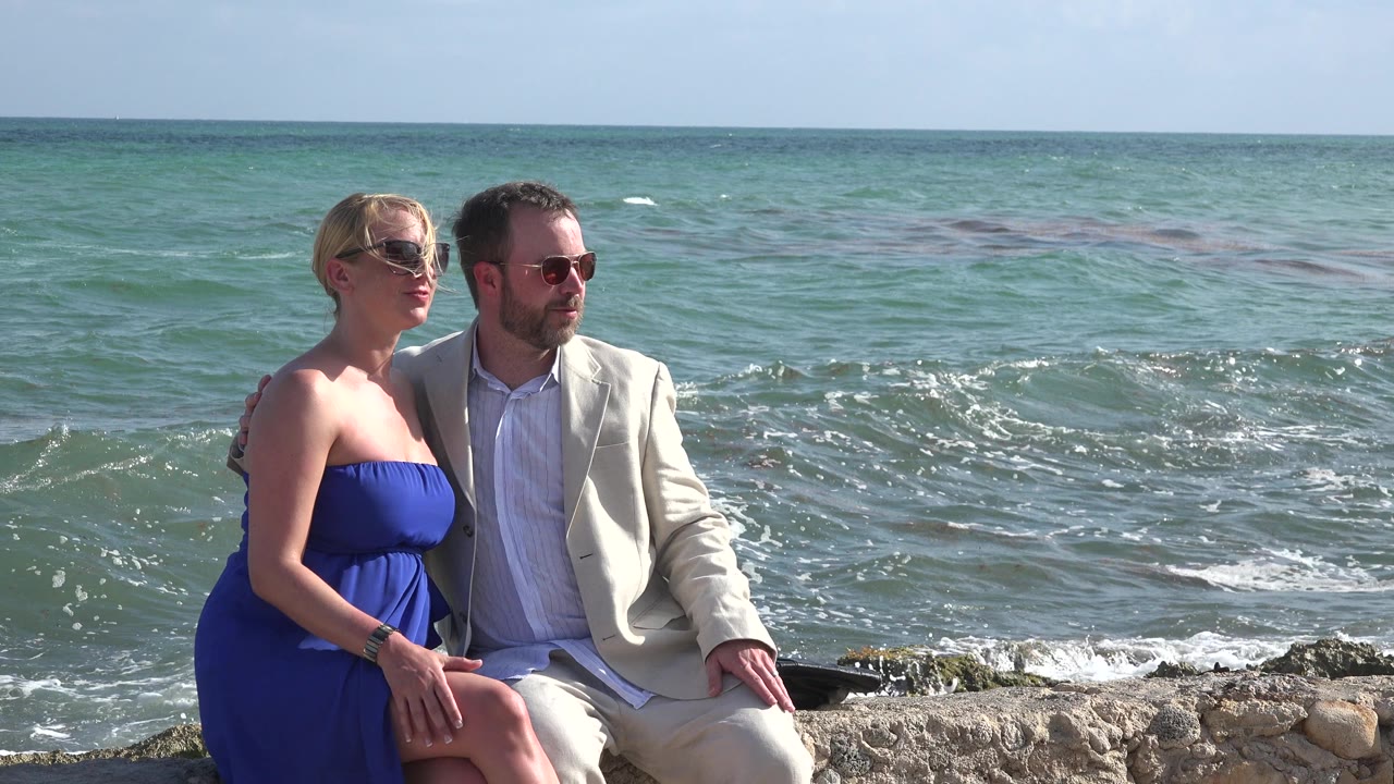 In Playa del Carmen, Mexico, a woman in a blue dress and a man in a light suit sit close together on rocks while waves roll behind them.