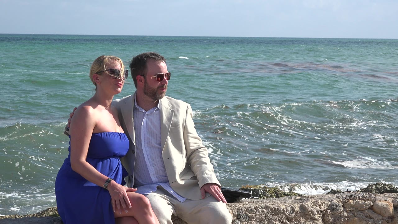 The couple remains seated on the low rocks in Playa del Carmen, Mexico, with the woman facing forward and the man looking out toward the sea.
