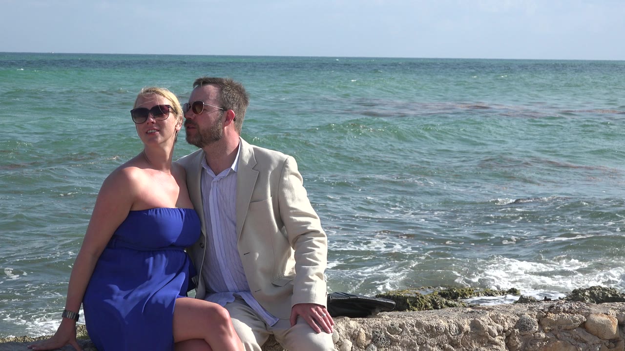 The couple remains seated on the stone shoreline in Playa del Carmen, Mexico, with the woman turned slightly left and the man looking toward her near the ocean horizon.