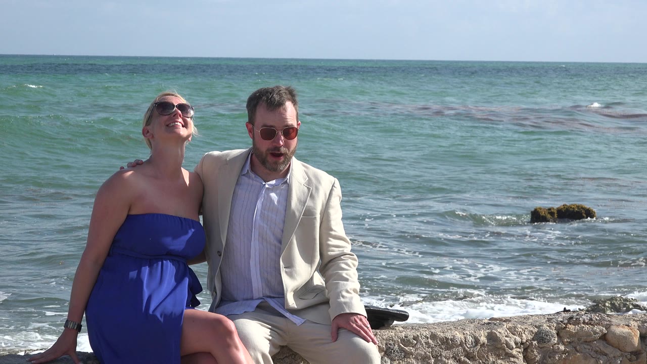 Couple in sunglasses seated on a stone wall beside the ocean