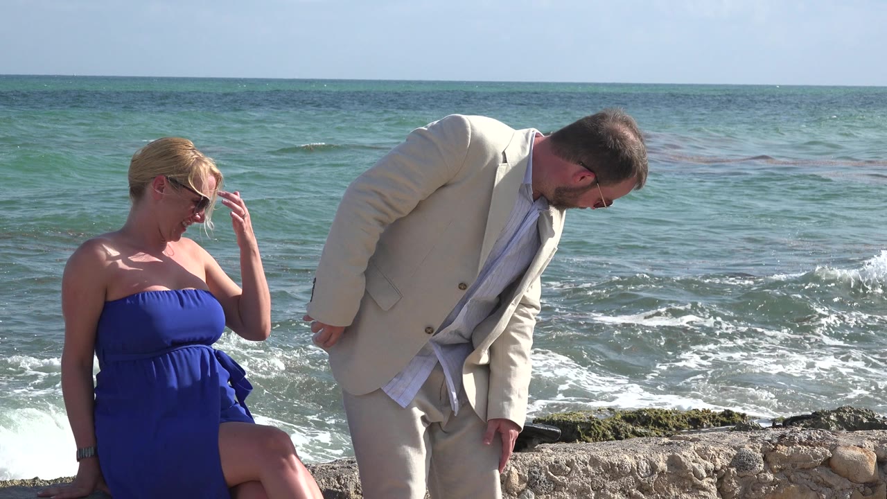 At Playa del Carmen, Mexico, a woman in a blue strapless dress sits on shoreline rocks while a man in a light suit leans nearby with the ocean behind them.
