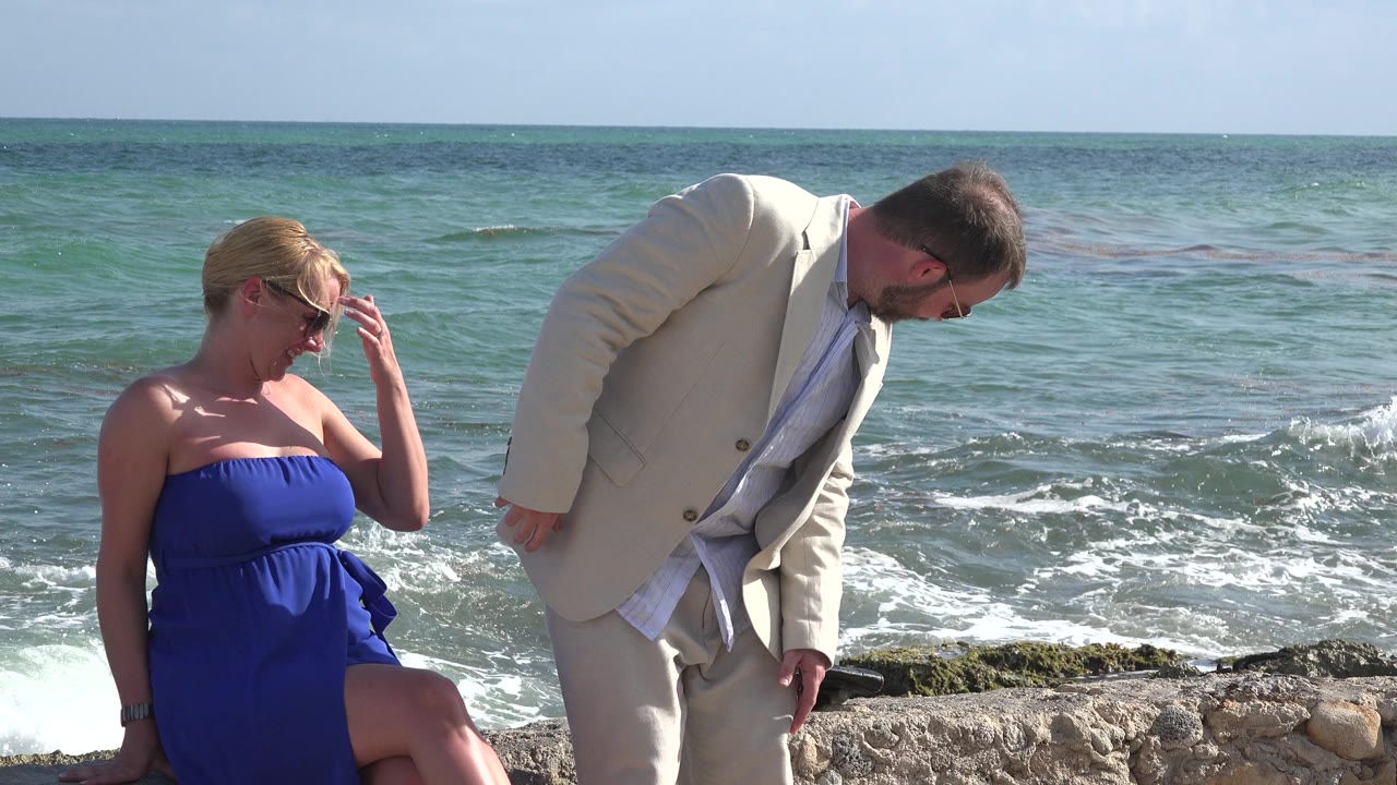 At Playa del Carmen, Mexico, the seated woman adjusts near her sunglasses while the man bends toward the rocks and foamy waves roll in behind them.