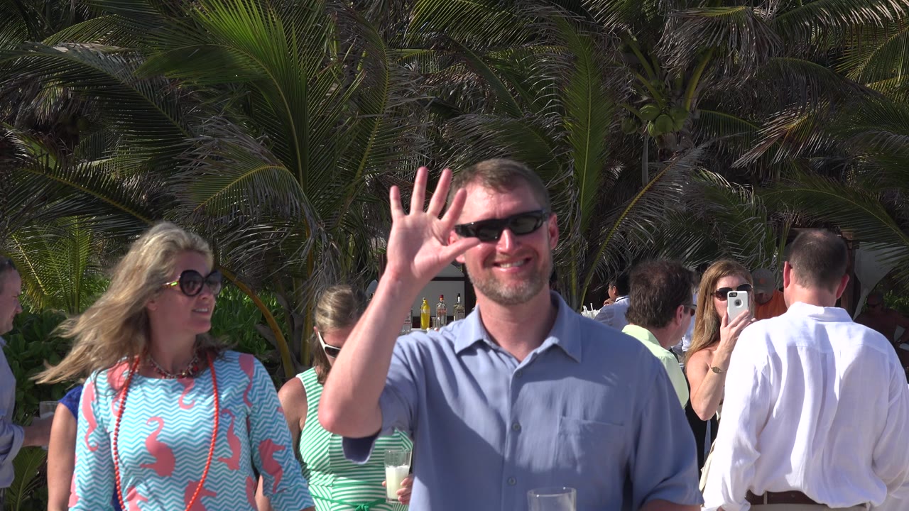 In Playa del Carmen, Mexico, a man in sunglasses waves toward the camera while a woman in a turquoise patterned dress stands beside him with palm fronds and guests behind them.