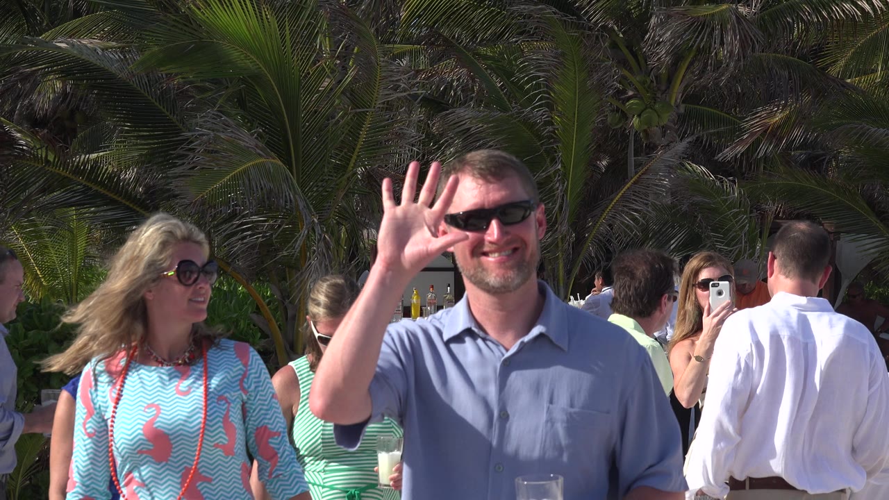 In Playa del Carmen, Mexico, the waving man remains near the center as a woman in sunglasses and several tourists in light shirts stand among palms and hedges.