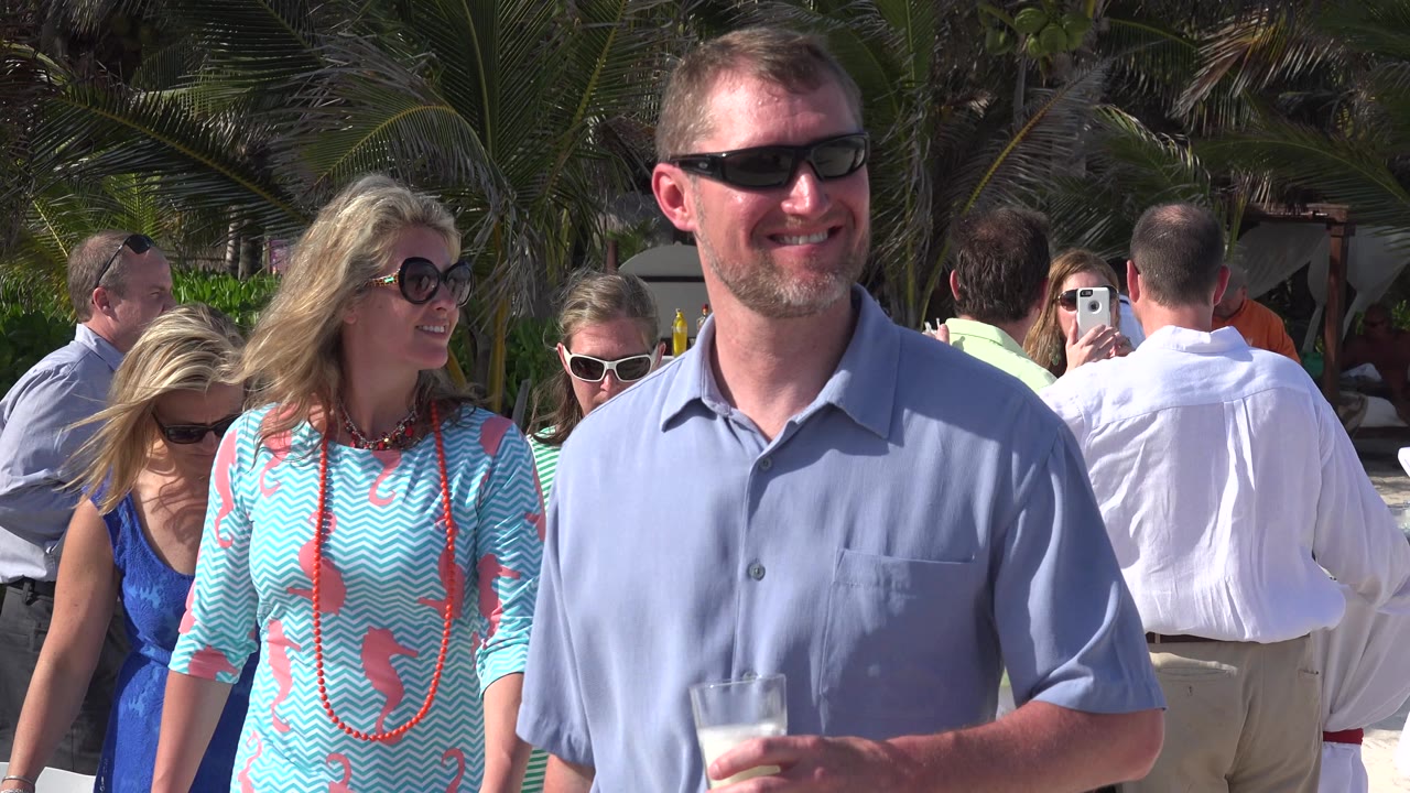 Man holding a drink while tourists stand under palm trees.