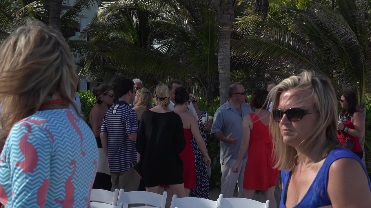 A woman in a blue dress and sunglasses stands in the foreground while adults mingle among white chairs and palms in Playa del Carmen, Mexico.