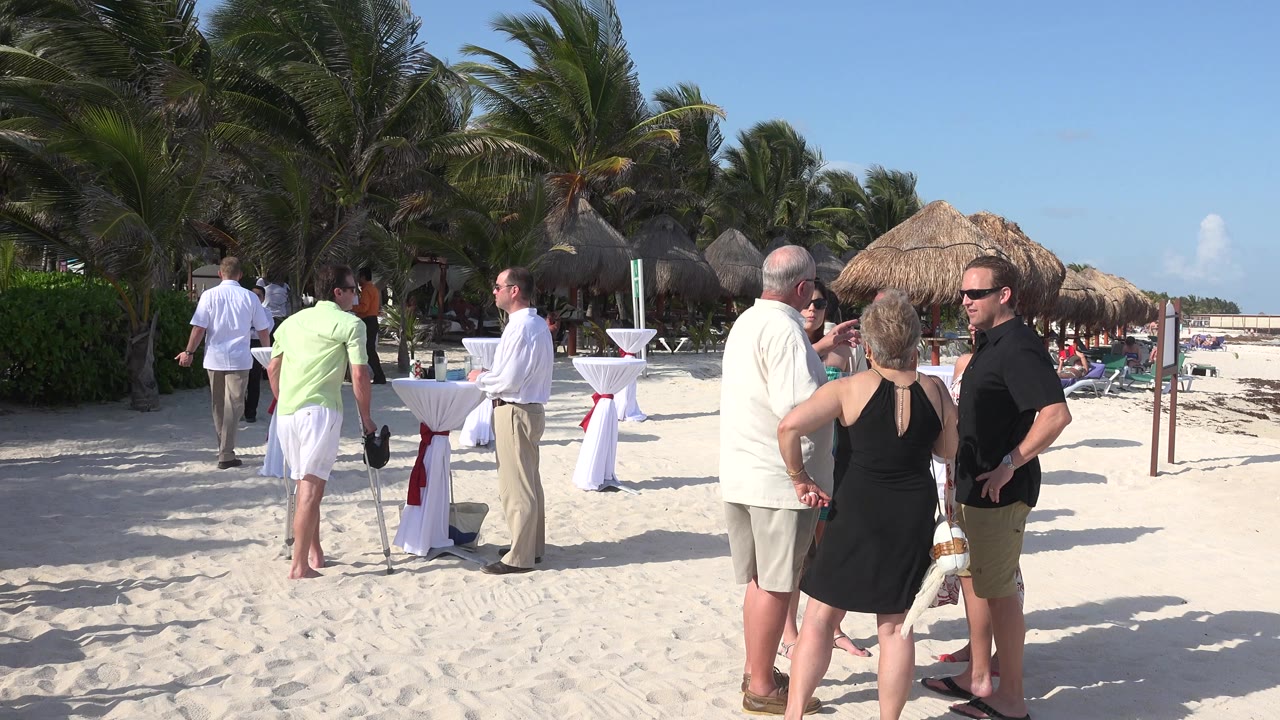 A small group of adults stands in the sand in Playa del Carmen, Mexico, with a man in white near cocktail tables and palms filling the shaded background.