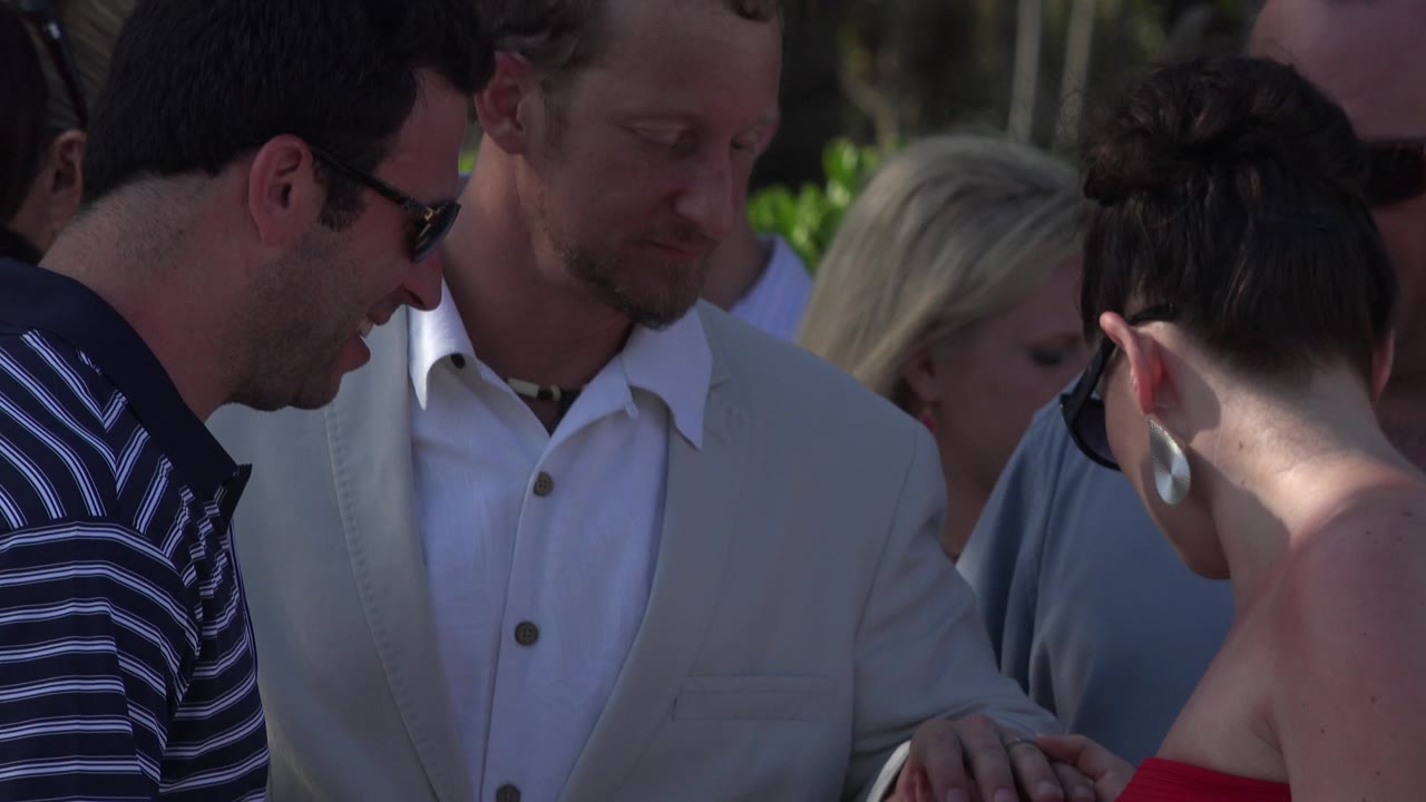 Adults stand close together in Playa del Carmen, Mexico, with a man in sunglasses leaning at left, a man in a pale jacket near center, and leafy shade behind them.