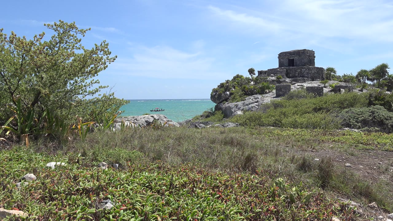 Opening frame in Tulum, Mexico shows a stone structure on a rocky rise above turquoise water, with low grass in front, a leafy tree at left, and no visible people.