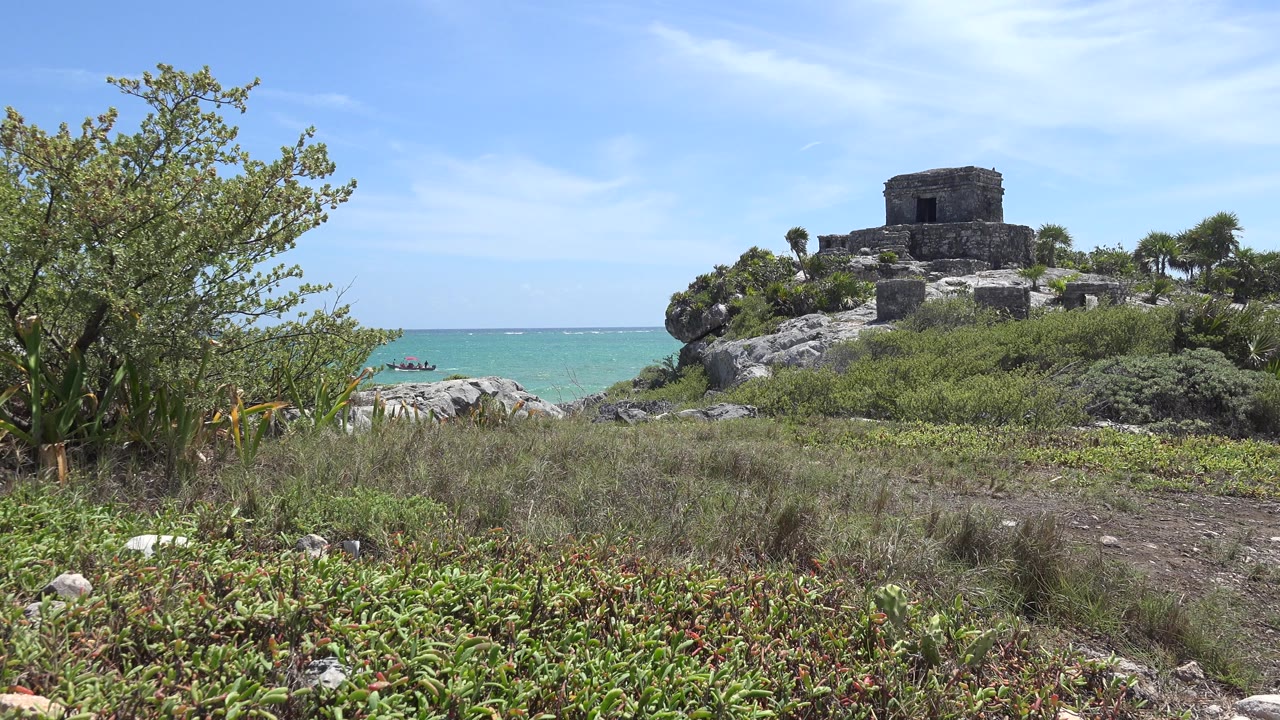 Opening frame of a Tulum, Mexico coastal view with foreground shrubs, a rocky rise, a small stone structure, turquoise water, blue sky, and no visible people.