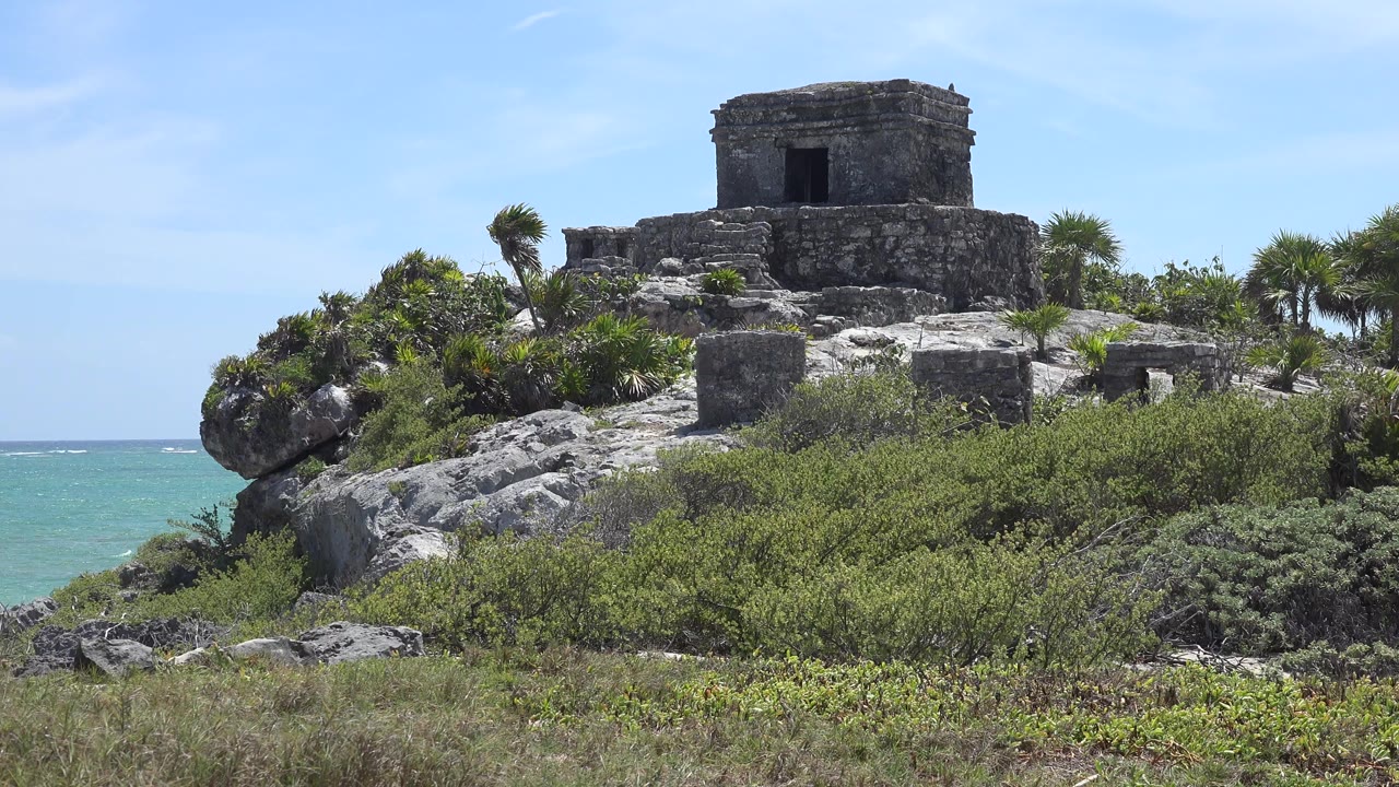 Opening frame of a stone fortress on a rocky hill in Tulum, Mexico, with green shrubs below, turquoise sea at the left edge, blue sky above, and no visible people.