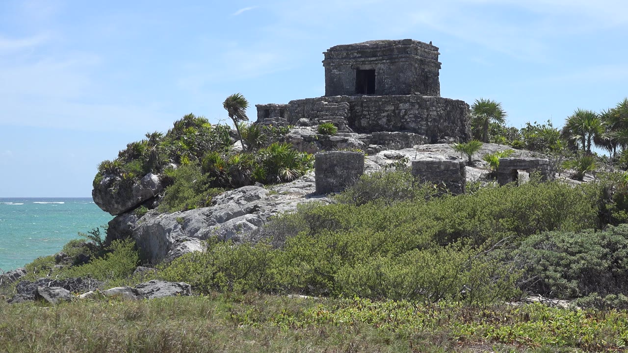 A stone temple in Tulum Mexico sits on a rocky rise above dense green shrubs, with turquoise ocean visible at left and no visible people.