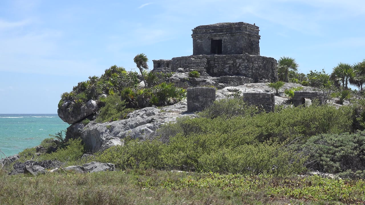 Stone ruin in Tulum, Mexico stands above rocky ground and dense coastal vegetation, with turquoise sea at left and no visible people.