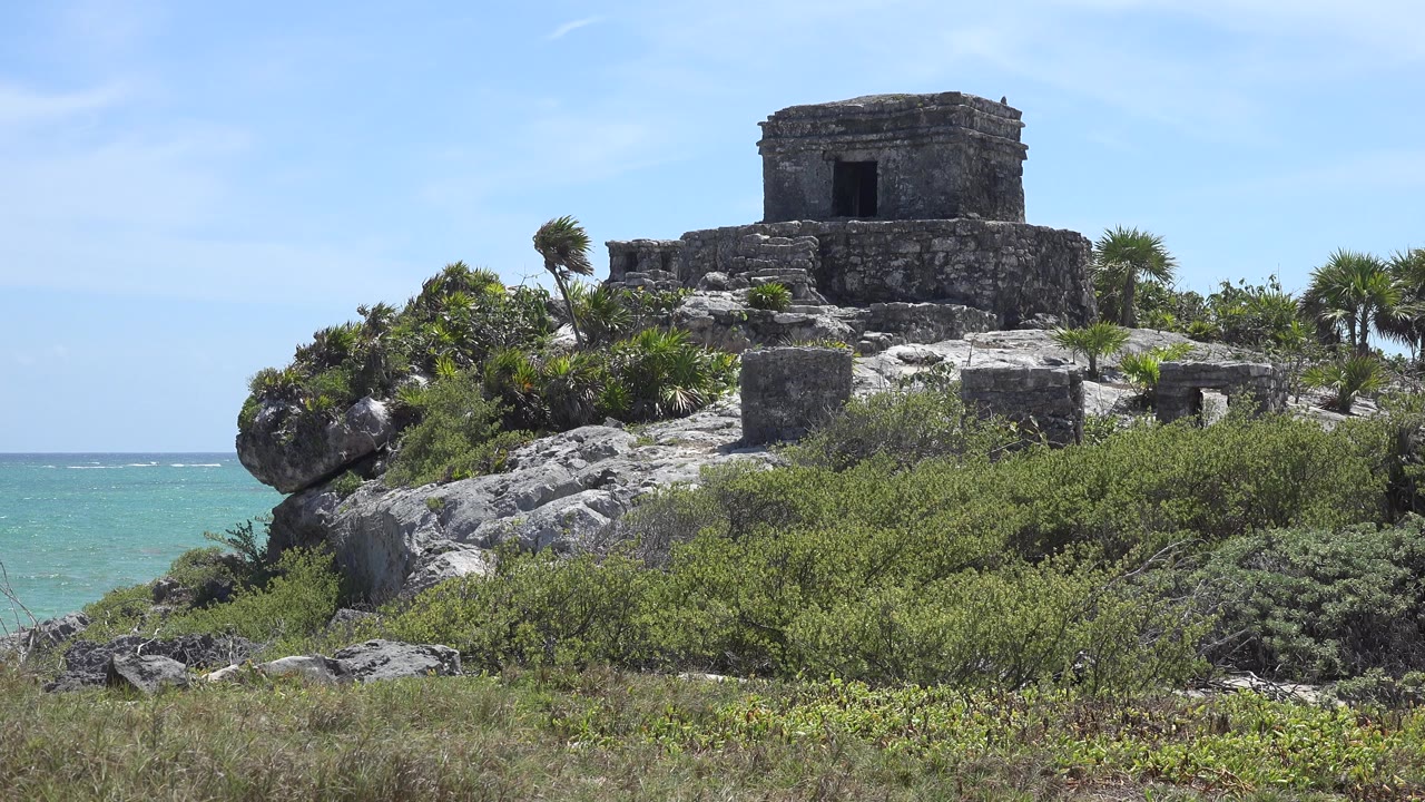At 0:01, the castle remains centered high in frame, with sunlit rocks and dense green shrubs below it in Tulum, Mexico, and no visible people.