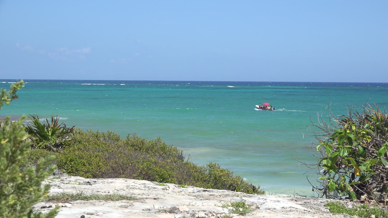 Opening frame of Tulum, Mexico shoreline with pale sand, green shrubs, turquoise water, a small red boat offshore, clear sky, and no visible people.