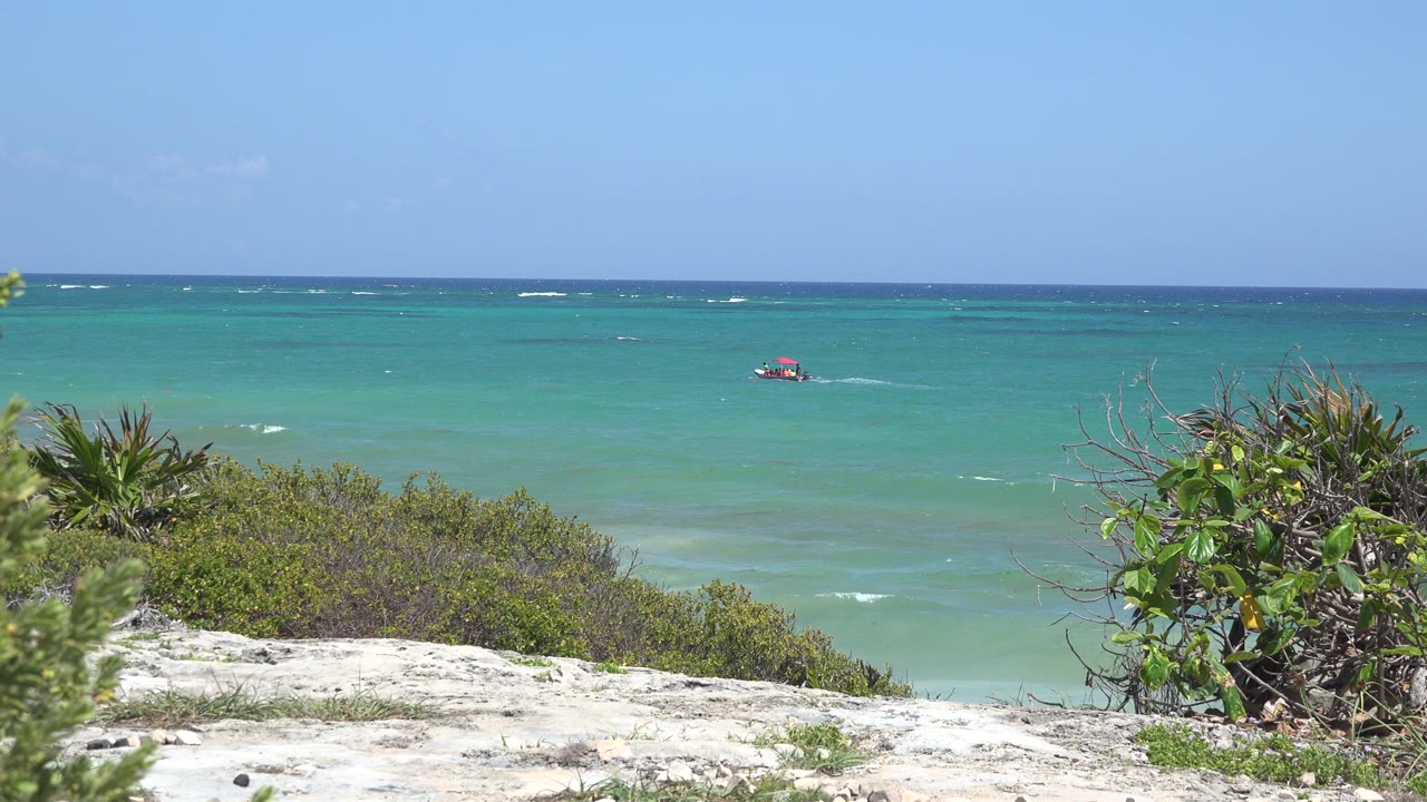 Opening frame in Tulum, Mexico, showing blue-green ocean, a small red object offshore, pale rock, low shrubs, spiky plants, clear sky, and no visible people.