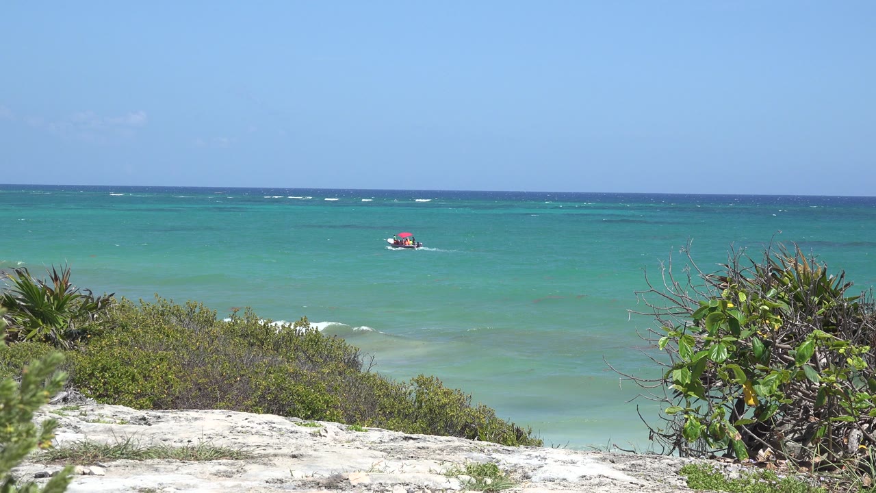 Turquoise ocean behind sandy beach shrubs with a small red boat offshore.