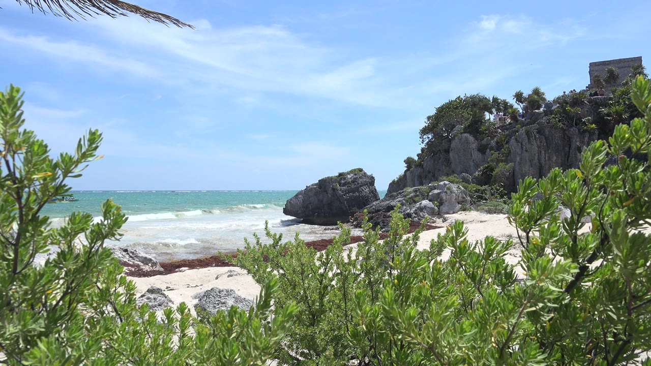 Green coastal shrubs frame rocks, beach, blue ocean, and a stone structure on a rocky rise.